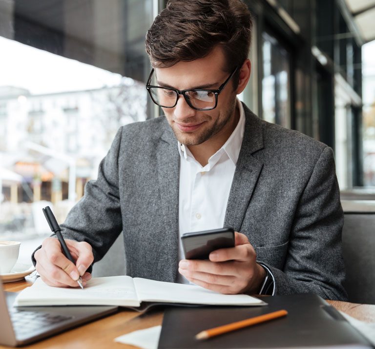 smiling-businessman-in-eyeglasses-sitting-by-the-table-in-cafe-with-laptop-computer-while-using-smartphone-and-writing-something-2